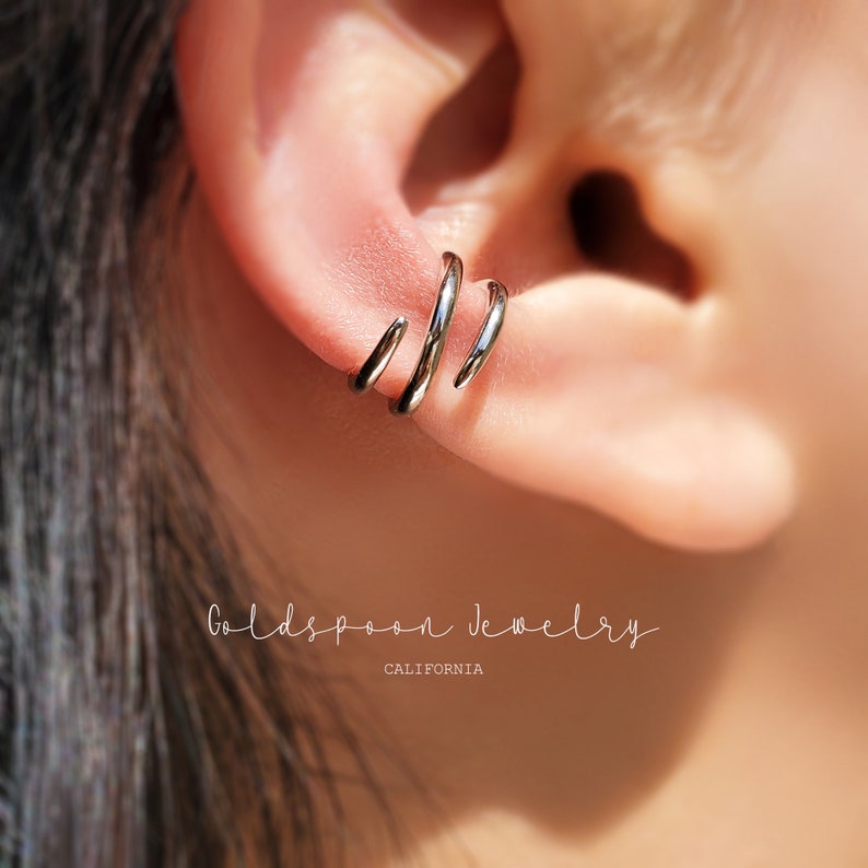 May include: Three silver hoop earrings worn in the helix piercing of an ear. The earrings are stacked on top of each other, with the largest hoop at the bottom and the smallest hoop at the top. The earrings are made of a smooth, shiny metal.