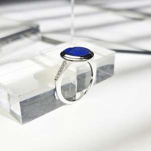 May include: A silver ring with a blue oval gemstone set in a bezel setting. The ring has a row of small clear gemstones on the band.