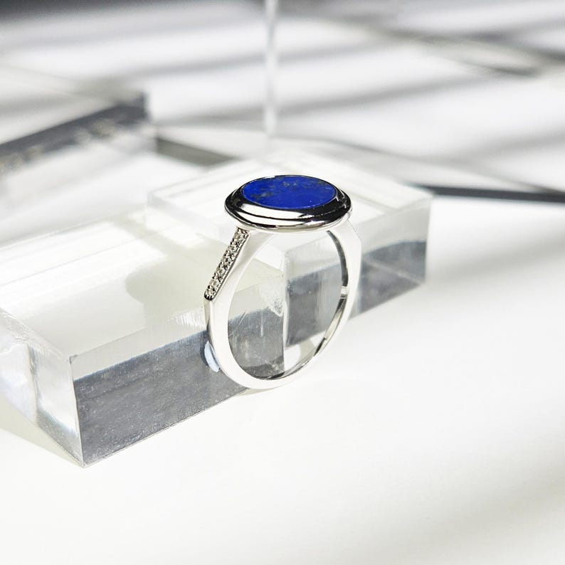 May include: A silver ring with a blue oval gemstone set in a bezel setting. The ring has a row of small clear gemstones on the band.