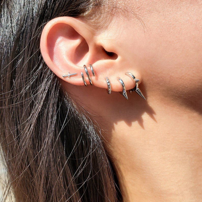 May include: A person's ear with multiple silver earrings. The earrings include a small dagger, two hoop earrings, and two spiked hoop earrings.
