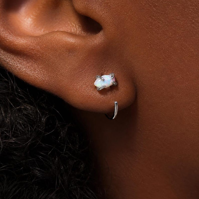 Puede incluir: Un piercing de oreja en tono plateado con una piedra de &oacute;palo iridiscente de corte marquesa sujeta por cuatro garras. La joya incluye una barra de metal curvada que rodea el l&oacute;bulo de la oreja, creando un aspecto elegante y moderno.