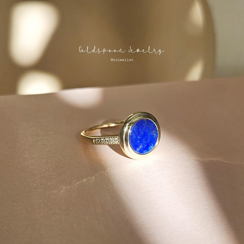 May include: A gold ring with a round blue gemstone and a row of small diamonds. The ring is on a pink surface. Goldspoon Jewelry Minimalist.