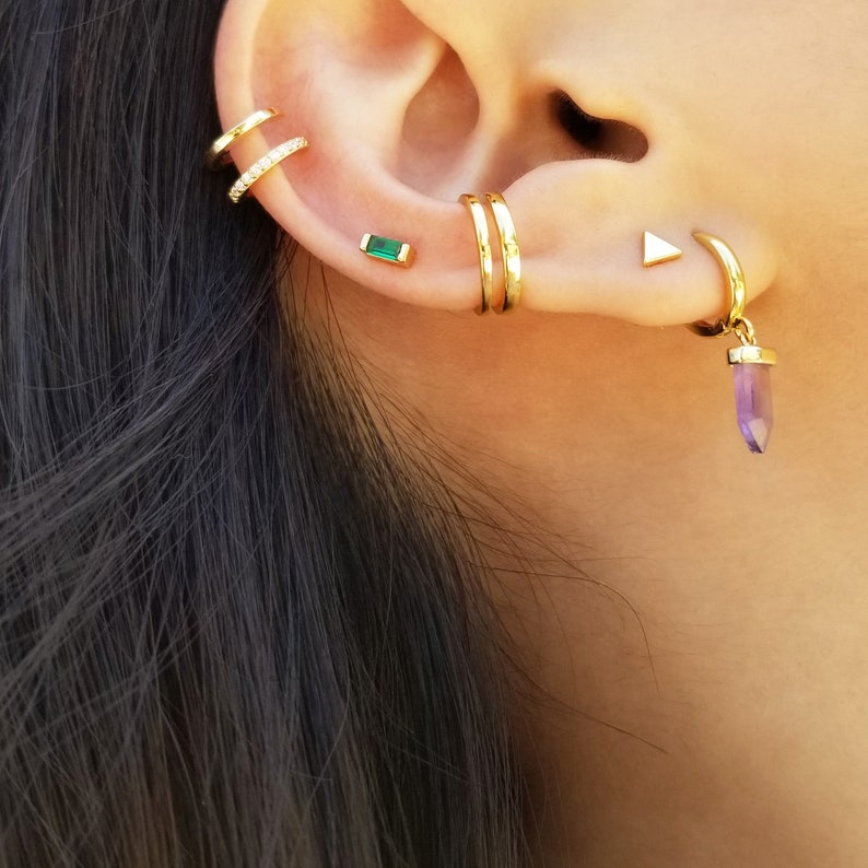May include: A woman's ear with multiple gold earrings. The earrings include a small emerald green stone, a triple hoop earring, a small triangle stud, and a hoop earring with a dangling amethyst stone.