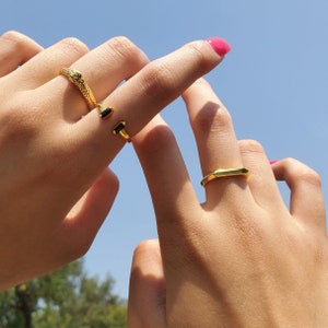 May include: Two gold rings on a person's fingers. One ring is a snake design with a black stone, the other is a simple rectangle shape.