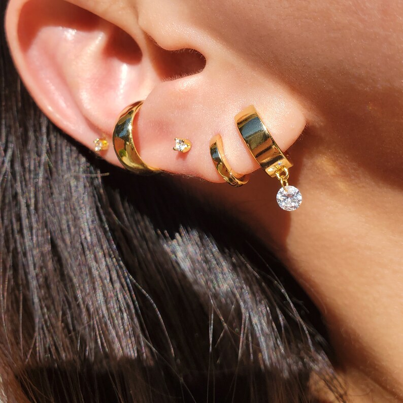 May include: Gold hoop earrings with a dangling clear crystal. The earrings are worn in the upper earlobe.