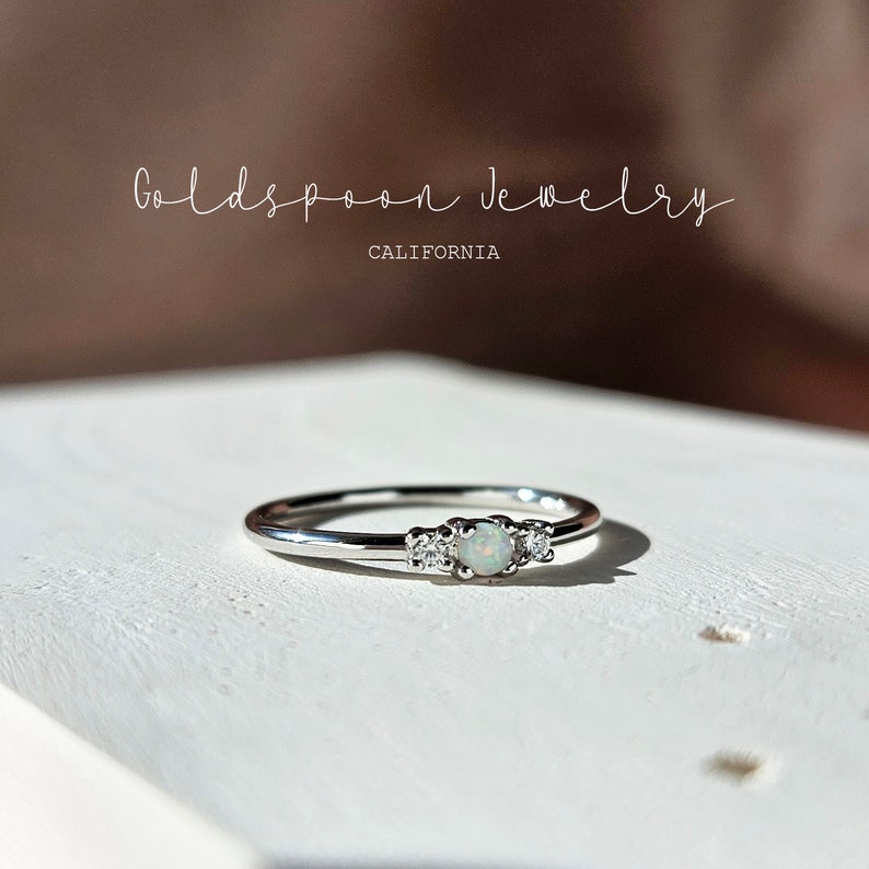 May include: A silver ring with a central opal stone flanked by two smaller clear stones. The ring is set against a white surface, with the text "Goldspoon Jewelry CALIFORNIA" in the background.