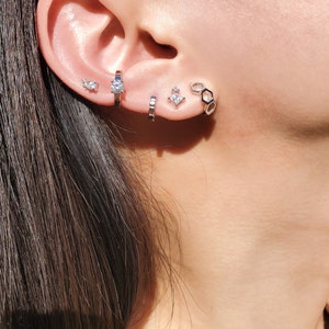 May include: A close-up of a person's ear with multiple silver earrings. The earrings include a small round stud, a larger round stud, a small geometric stud, and a larger hexagonal hoop.