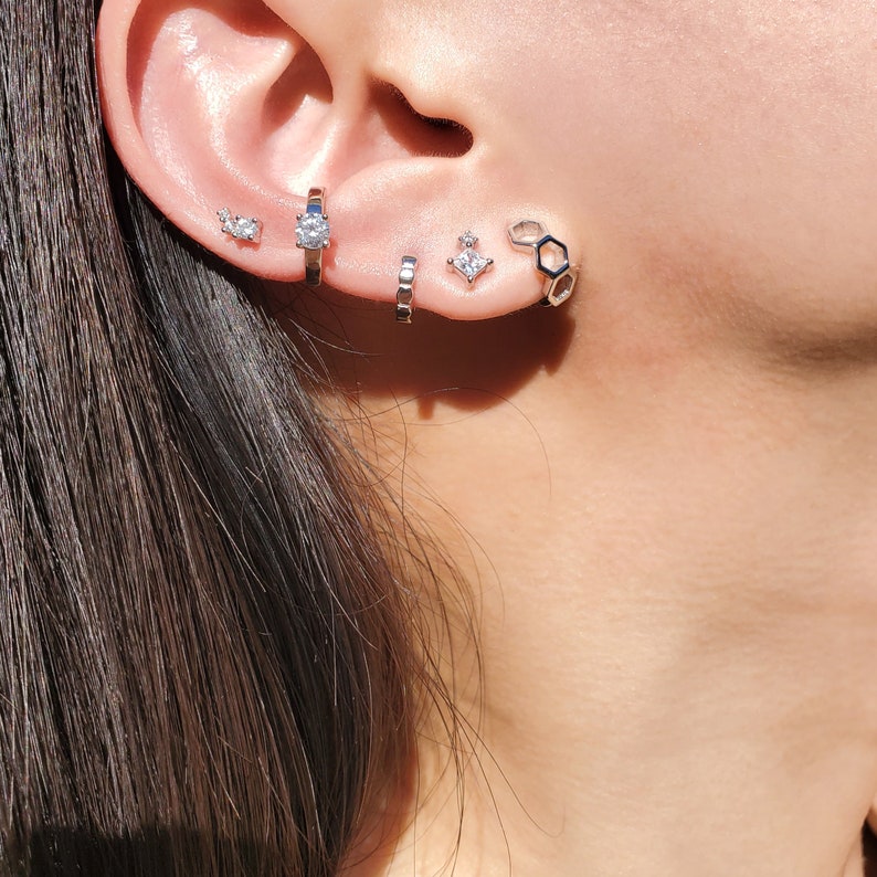May include: A close-up of a person's ear with multiple silver earrings. The earrings include a small round stud, a larger round stud, a small geometric stud, and a larger hexagonal hoop.