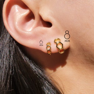 May include: Two gold honeycomb hoop earrings, one 6mm and one 8mm in diameter. The earrings are shown on a person's ear.