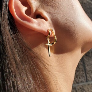 K&ouml;nnte beinhalten: Goldene Creolen mit einem baumelnden Kreuzanh&auml;nger. Der Creolen ist einfach und gebogen, das Kreuz ist ein klassisches Design.