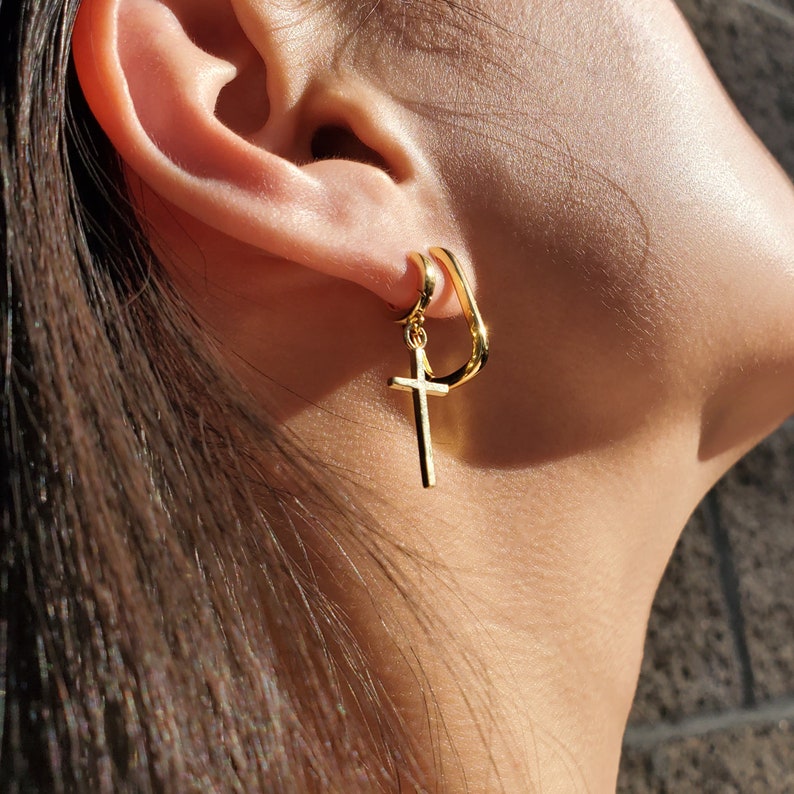 K&ouml;nnte beinhalten: Goldene Creolen mit einem baumelnden Kreuzanh&auml;nger. Der Creolen ist einfach und gebogen, das Kreuz ist ein klassisches Design.