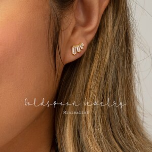 May include: Gold earrings with a row of small, clear, rectangular stones set in a curved shape. The earrings are shown on a person's ear.