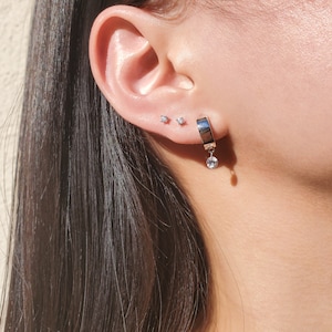 May include: A silver hoop earring with a dangling crystal charm. The earring is worn in the upper lobe of the ear. Two smaller silver stud earrings are also worn in the ear.