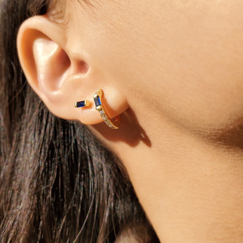 May include: A pair of gold earrings with a blue gemstone. The larger earring is a hoop with a row of clear gemstones. The smaller earring is a simple bar with a blue gemstone.
