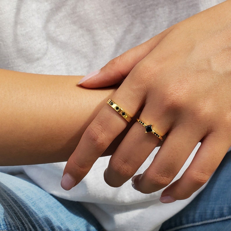 Op de afbeelding: Twee gouden ringen met zwarte edelstenen. De eerste ring is een dunne band met vijf kleine zwarte edelstenen. De tweede ring heeft een grotere vierkante zwarte edelsteen in het midden met kleinere zwarte edelstenen eromheen.