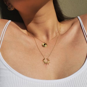 May include: A layered gold necklace set. The top necklace features a circular pendant with a black center. The bottom necklace has a sunburst pendant. The jewelry is worn over a white tank top.