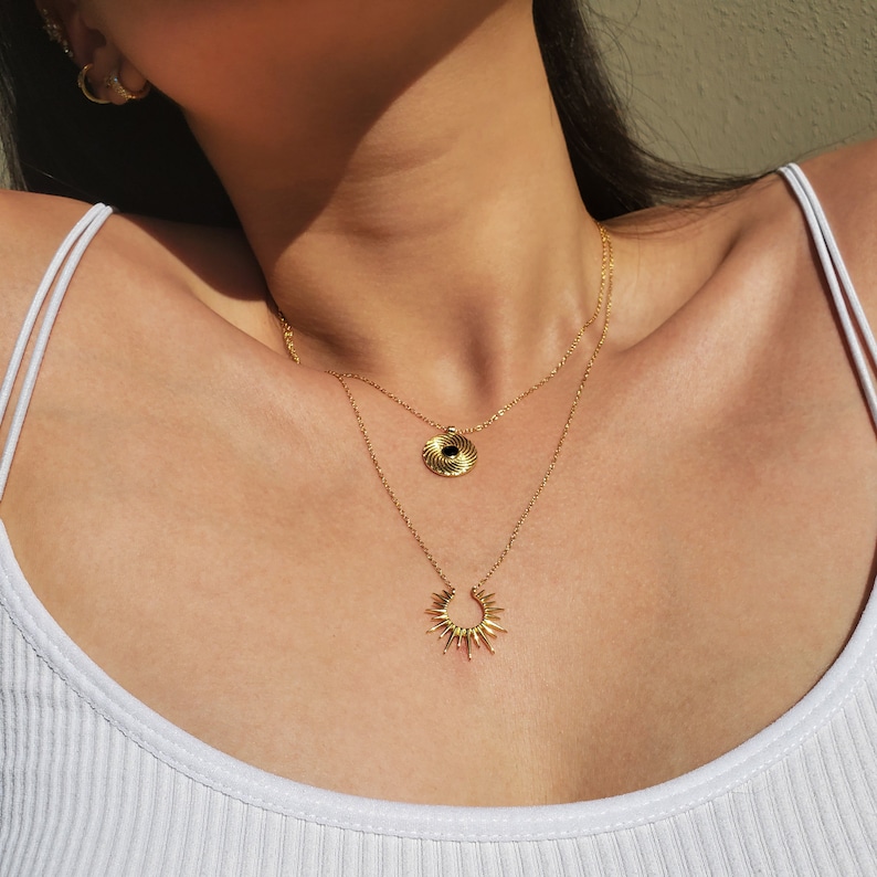 May include: A layered gold necklace set. The top necklace features a circular pendant with a black center. The bottom necklace has a sunburst pendant. The jewelry is worn over a white tank top.