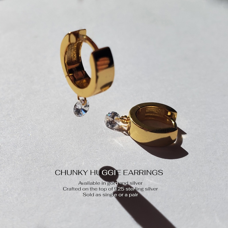 May include: Gold chunky huggie earrings with a small clear stone dangling from each earring.  "CHUNKY HUGGIE EARRINGS" text is below the earrings.  "Available in gold and silver"  "Crafted on the top of 925 sterling silver"  "Sold as single or a pair"