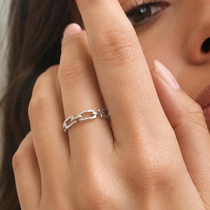 May include: A silver-toned ring featuring a continuous chain-link design. The band is composed of small, rectangular metal links connected in a repeating pattern, creating a polished and uniform appearance around the entire circumference of the ring.