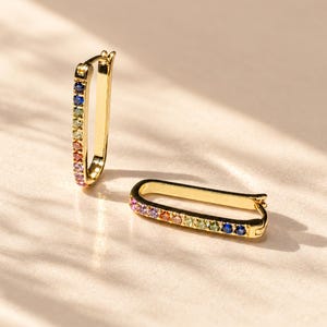 May include: A pair of gold-toned hoop earrings with an elongated, rectangular shape. Each earring features a row of small, multi-colored gemstones set along one side, displaying a gradient of blue, green, orange, and purple hues.