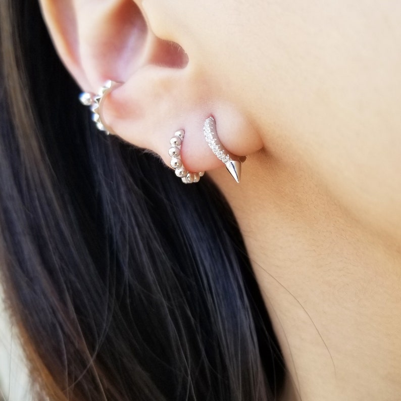 May include: A close-up of a person's ear with three silver earrings. The bottom earring is a small hoop with a pointed tip. The middle earring is a small hoop with a row of small stones. The top earring is a cluster of small silver beads.