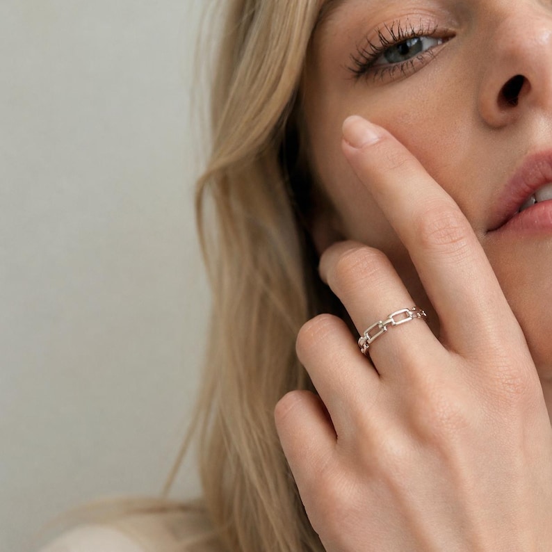 May include: A silver-toned ring featuring a delicate chain-link design. The band is composed of small, rectangular metal links connected in a continuous loop, creating a textured appearance that encircles the finger with a polished finish.