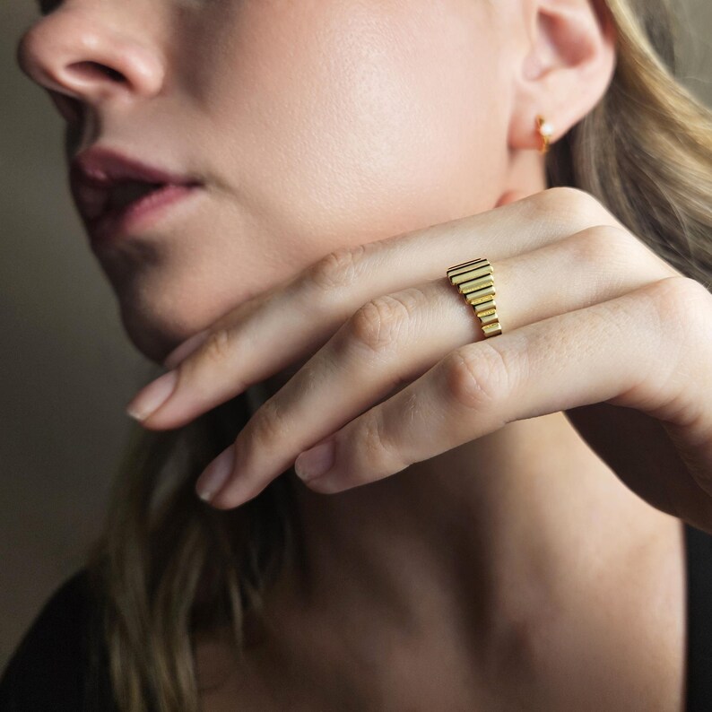 May include: A gold ring with a textured, stepped design. The ring is worn on a person's finger.