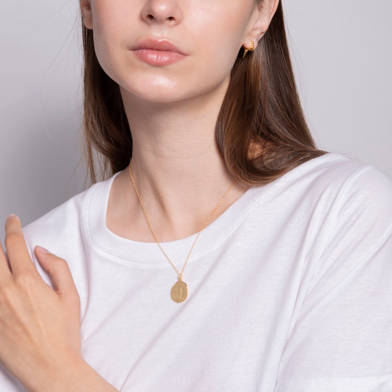 May include: A gold necklace with a round pendant. The pendant has a textured surface. The necklace is worn by a person with long brown hair.
