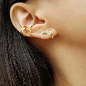 May include: Gold hoop earrings with a small green emerald stone stud earring. The earrings are worn in the upper earlobe and helix piercing.