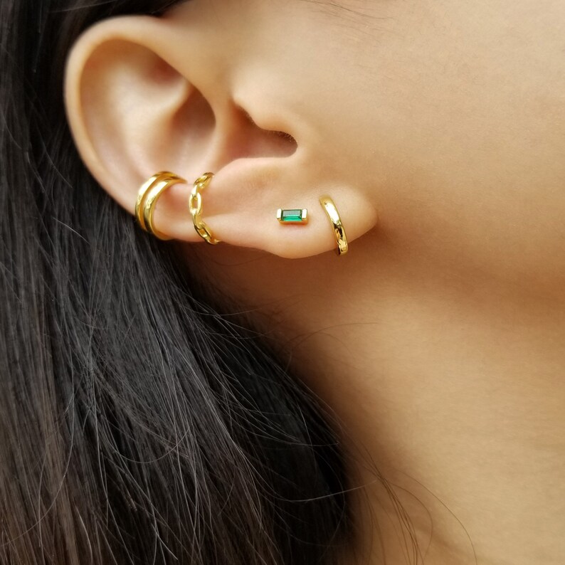May include: Gold hoop earrings with a small green emerald stone stud earring. The earrings are worn in the upper earlobe and helix piercing.