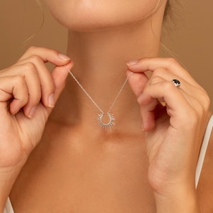 May include: A silver necklace with a sunburst pendant. The necklace is held up by a person's hands. A ring with a dark stone is on the person's finger. The background is a solid tan color.