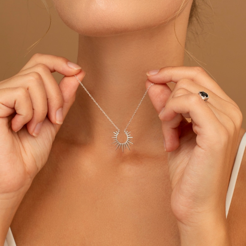 May include: A silver necklace with a sunburst pendant. The necklace is held up by a person's hands. A ring with a dark stone is on the person's finger. The background is a solid tan color.