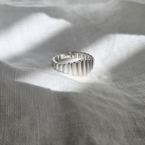 May include: A silver ring with a unique, textured design. The ring is made of multiple vertical bars that create a wave-like pattern.