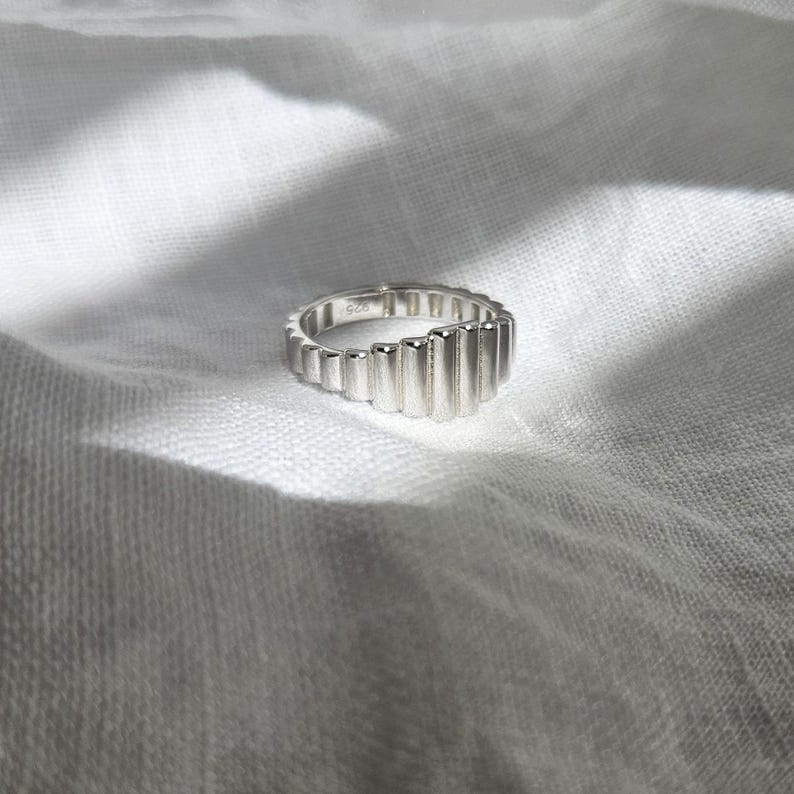 May include: A silver ring with a unique, textured design. The ring is made of multiple vertical bars that create a wave-like pattern.