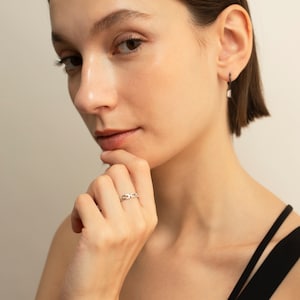 May include: A gold-coloured chain link ring is worn on a person's finger. The ring is made of small, rectangular links. The person is wearing a black top and has dark hair. The background is a neutral colour.