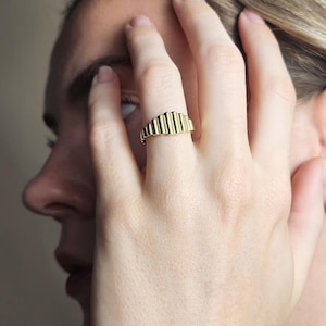 May include: A gold ring with a textured, ridged design. The ring is worn on a person's finger.