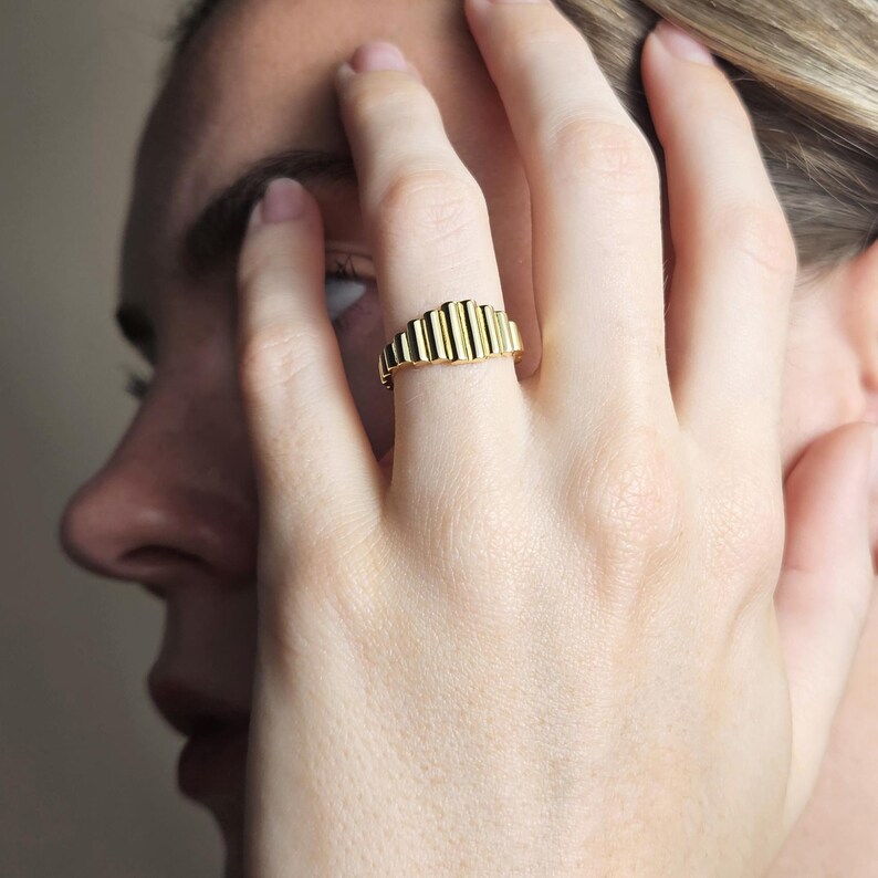 May include: A gold ring with a textured, ridged design. The ring is worn on a person's finger.