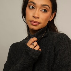 以下が含まれることがあります： 黒いタートルネックセーターを着た女性。金のチェーンネックレスと黒い石の金の指輪をしている。