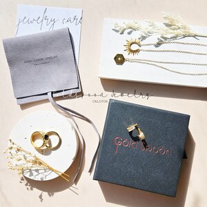 May include: Gold Spoon Jewelry packaging with a gray velvet pouch, a white stone dish with two gold rings, and a black box with gold lettering. The jewelry includes a gold necklace with a sun pendant and a hexagon pendant.