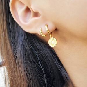 May include: Gold hoop earrings with a small beaded hoop and a larger hoop with a dangling charm featuring a sun design.
