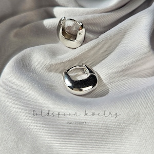 May include: Two silver hoop earrings on a white fabric background. The earrings are simple and modern, with a smooth, rounded shape. The text "Goldspoon Jewelry California" is visible in the bottom center of the image.