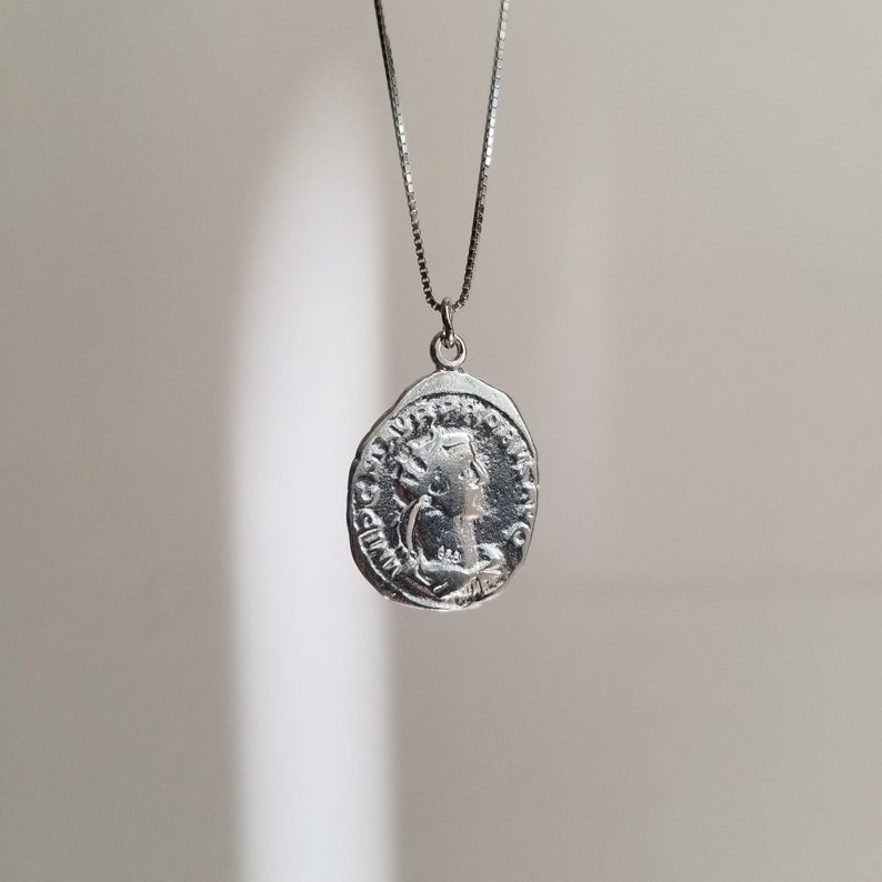 May include: Silver necklace with a round coin pendant featuring a profile of a woman with a crown. The coin has Latin lettering around the edge.