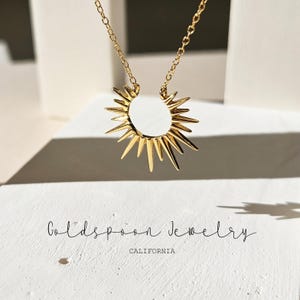 以下が含まれることがあります： ゴールドのネックレスに、太陽の光を思わせるペンダントが飾られています。ペンダントはゴールドメタル製で、円形に鋭いポイントが外側に放射状に伸びています。ネックレスは白い背景に置かれ、その下に「Goldspoon Jewelry California」の文字が書かれています。