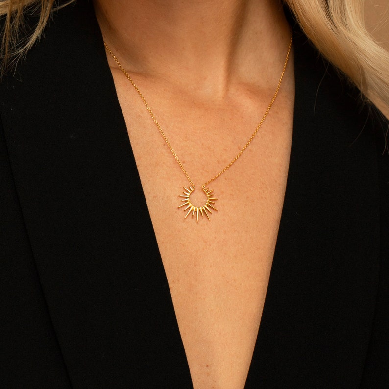 May include: A gold-colored necklace featuring a sunburst pendant. The pendant has a series of pointed rays emanating from a central point. The necklace is worn against a black garment, creating a contrast that highlights the jewelry.