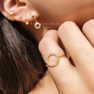 May include: A gold ring with a circle of small clear stones. The ring is on a person's finger.