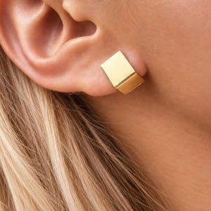 Puede incluir: Un ear cuff dorado con un diseño de placa metálica rectangular y lisa que se ajusta al borde exterior del cartílago de la oreja. La pieza presenta una superficie pulida y reflectante con bordes limpios y una construcción geométrica sólida.