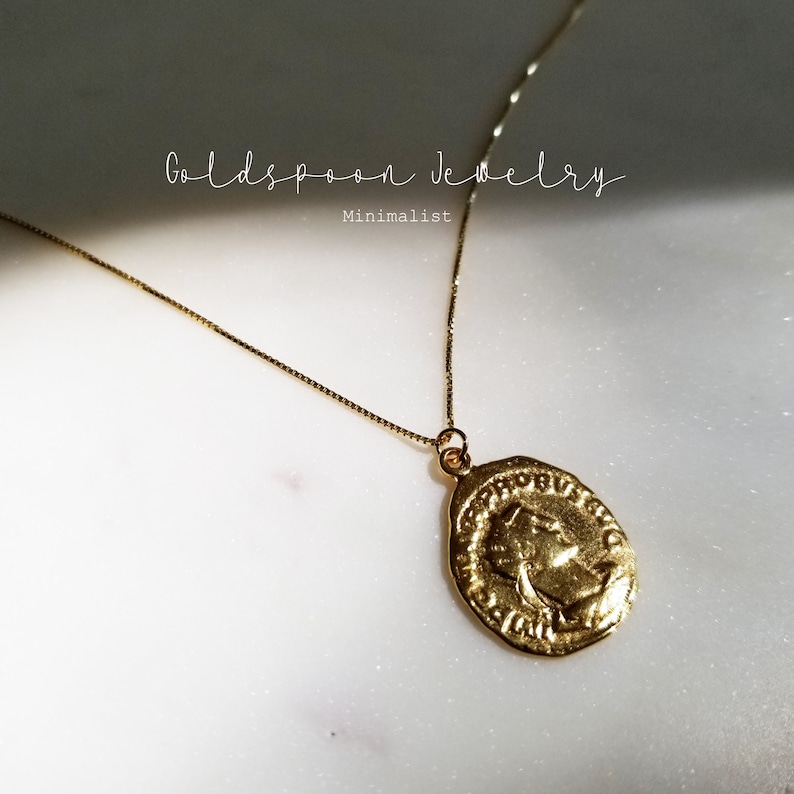 May include: Gold chain necklace with a gold oval pendant featuring a profile of a person's face. The pendant has an inscription around the edge. The text "Goldspoon Jewelry Minimalist" is visible in the background.