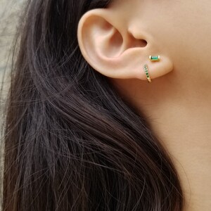 May include: A pair of gold earrings with green emerald stones. One earring is a hoop with a row of emerald stones, the other is a single emerald stone.