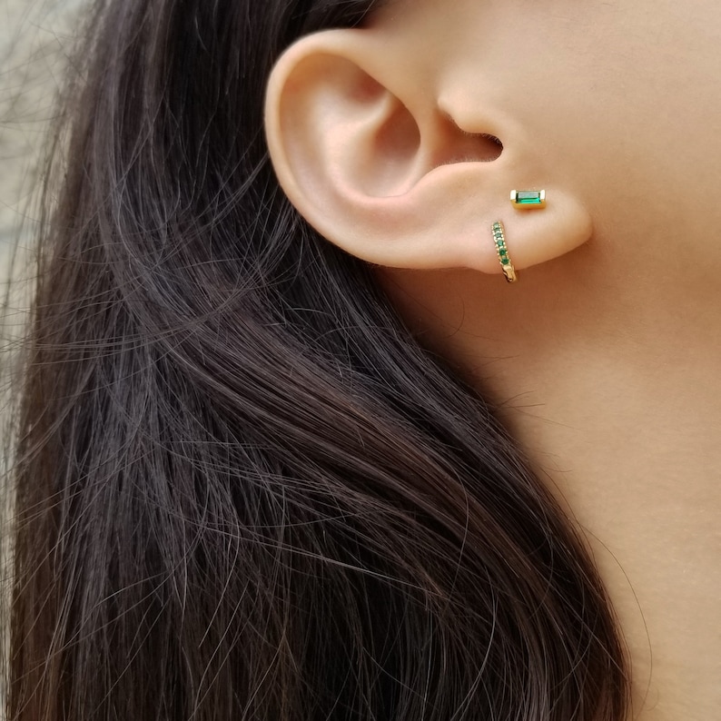 May include: A pair of gold earrings with green emerald stones. One earring is a hoop with a row of emerald stones, the other is a single emerald stone.