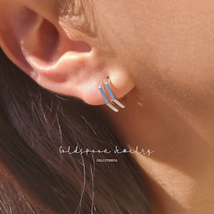 May include: A pair of silver bar earrings with a minimalist design. The earrings are worn in the earlobe of a person with a light complexion.